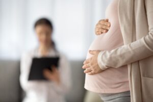 Pregnant woman stands with her hands on her stomach while asking her doctor, who sits nearby, “What constitutes medical negligence during childbirth?”
