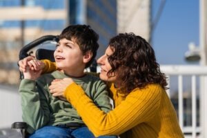 A mother embracing a boy with cerebral palsy sitting in a chair. Find out whether cerebral palsy affects your child’s lifespan.